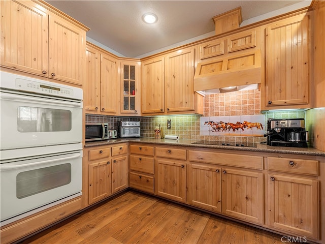 kitchen with black electric stovetop, backsplash, white double oven, dark stone counters, and light hardwood / wood-style flooring