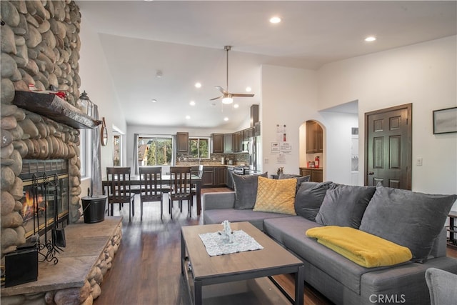 living room featuring a stone fireplace, high vaulted ceiling, dark wood-type flooring, and ceiling fan