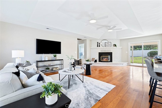 living room with hardwood / wood-style floors, ceiling fan, a brick fireplace, and a raised ceiling