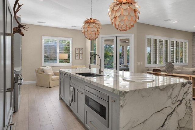 kitchen featuring sink, gray cabinets, appliances with stainless steel finishes, a kitchen island with sink, and light stone counters