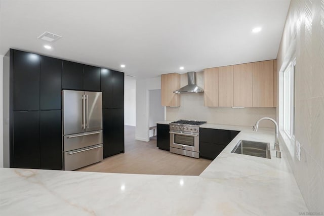 kitchen featuring high quality appliances, wall chimney range hood, sink, light brown cabinetry, and light hardwood / wood-style floors