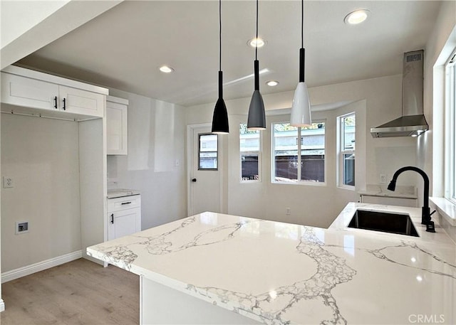 kitchen with wall chimney range hood, sink, hanging light fixtures, light stone counters, and white cabinetry