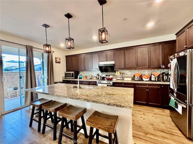 kitchen with stainless steel appliances, sink, decorative light fixtures, light hardwood / wood-style flooring, and an island with sink