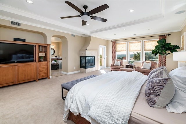 carpeted bedroom with ornamental molding, a large fireplace, a tray ceiling, ceiling fan, and connected bathroom