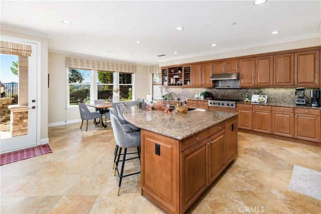 kitchen featuring light stone countertops, decorative backsplash, stainless steel gas cooktop, a kitchen island with sink, and crown molding