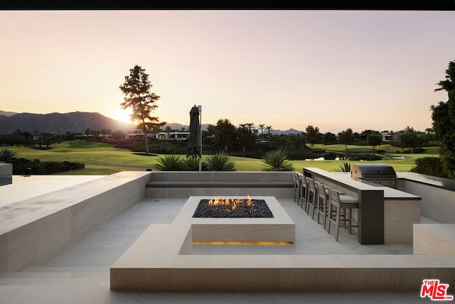 patio terrace at dusk featuring exterior kitchen, grilling area, an outdoor fire pit, a mountain view, and an outdoor bar