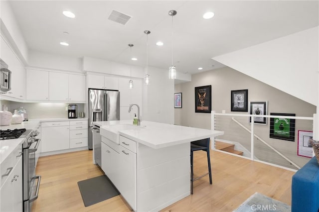 kitchen featuring stainless steel appliances, an island with sink, decorative light fixtures, white cabinets, and light wood-type flooring