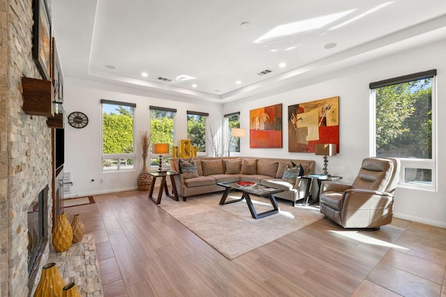 living room featuring a tray ceiling, a stone fireplace, a wealth of natural light, and light hardwood / wood-style floors