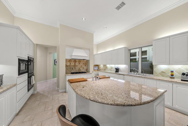 kitchen featuring light stone countertops, backsplash, an island with sink, white cabinets, and custom range hood