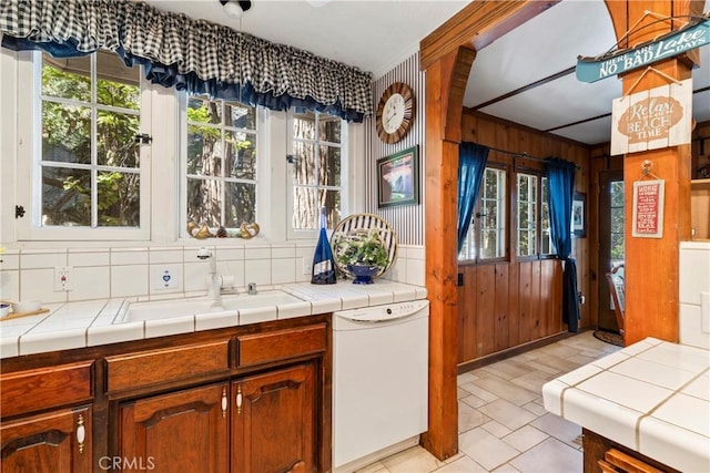 kitchen with a wealth of natural light, wooden walls, tile counters, and white dishwasher