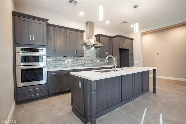 kitchen featuring decorative backsplash, double oven, wall chimney range hood, decorative light fixtures, and an island with sink