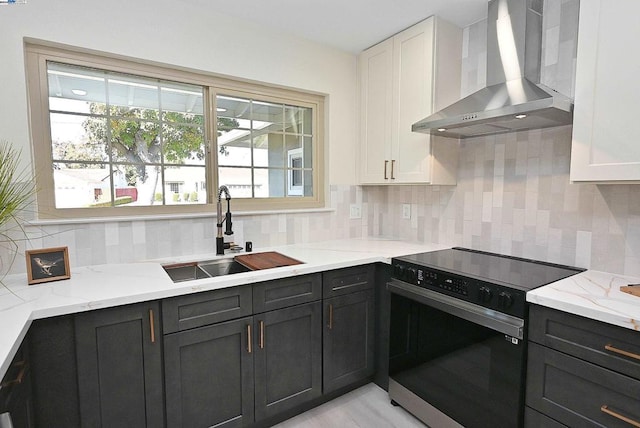 kitchen with light stone countertops, wall chimney range hood, electric range, white cabinetry, and sink