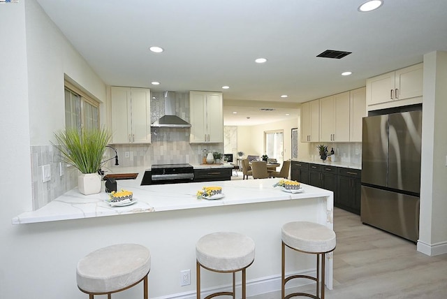 kitchen with kitchen peninsula, wall chimney exhaust hood, stainless steel appliances, a kitchen bar, and backsplash