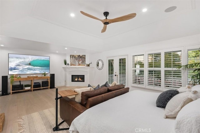 bedroom featuring light wood finished floors, a glass covered fireplace, ceiling fan, a tray ceiling, and recessed lighting