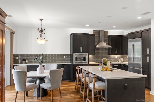 kitchen with wall chimney exhaust hood, built in appliances, decorative light fixtures, light hardwood / wood-style floors, and an island with sink