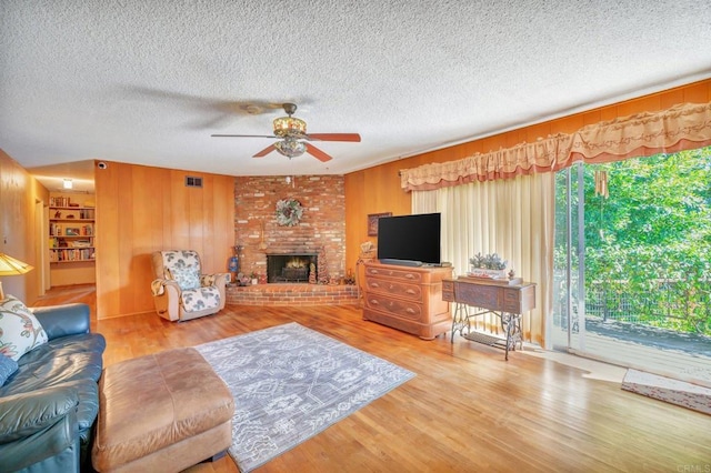 living room with wooden walls, wood-type flooring, ceiling fan, a brick fireplace, and a textured ceiling