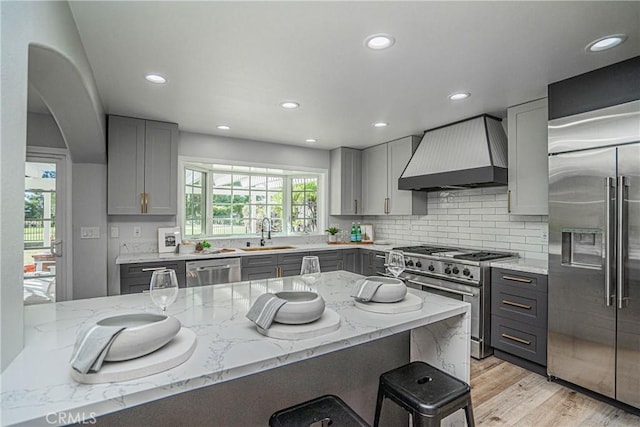 kitchen featuring light stone countertops, premium range hood, gray cabinets, high quality appliances, and light wood-type flooring