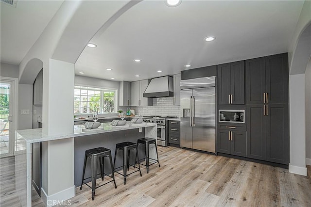 kitchen featuring light stone counters, wall chimney exhaust hood, built in appliances, light hardwood / wood-style floors, and a breakfast bar area