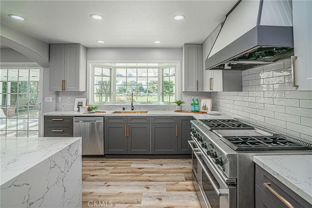 kitchen featuring gray cabinetry, light stone counters, premium range hood, appliances with stainless steel finishes, and light wood-type flooring