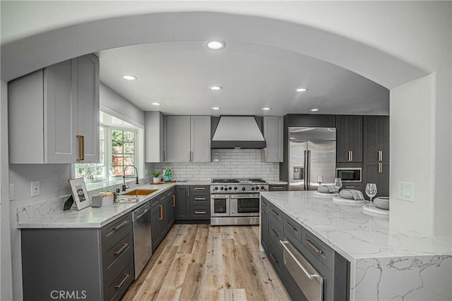 kitchen with gray cabinetry, wall chimney range hood, sink, premium appliances, and light hardwood / wood-style floors