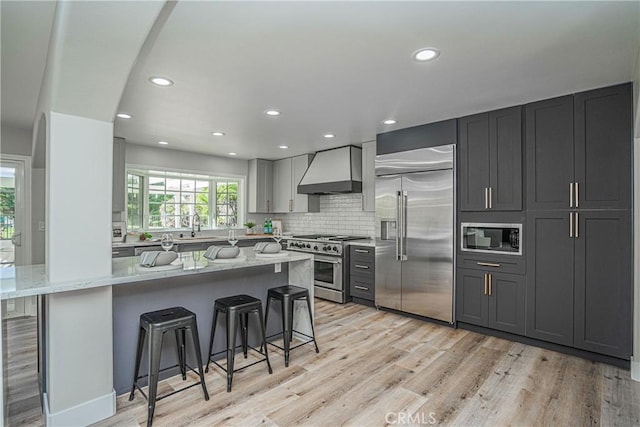 kitchen featuring a kitchen breakfast bar, light wood-type flooring, light stone counters, wall chimney range hood, and built in appliances