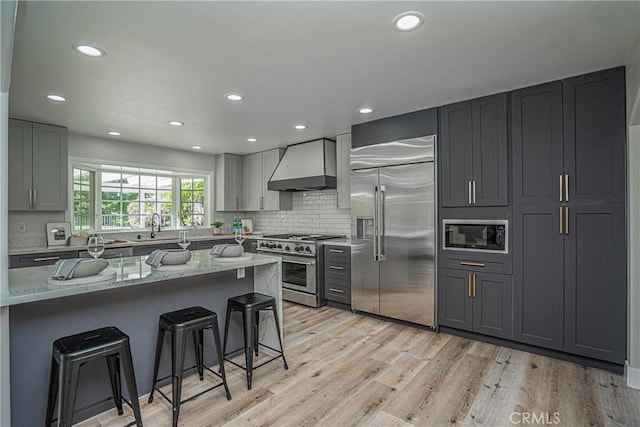 kitchen featuring light hardwood / wood-style floors, wall chimney range hood, built in appliances, gray cabinets, and a breakfast bar area