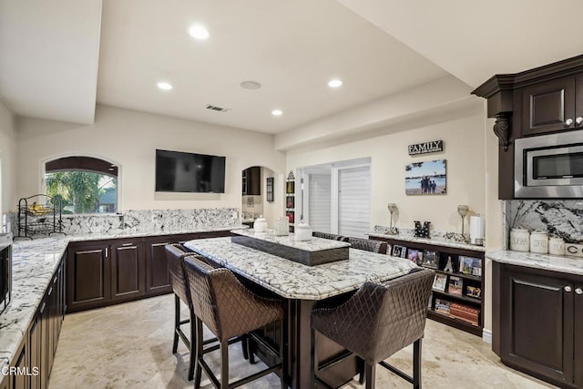 kitchen featuring dark brown cabinetry, stainless steel microwave, a kitchen breakfast bar, light stone counters, and decorative backsplash