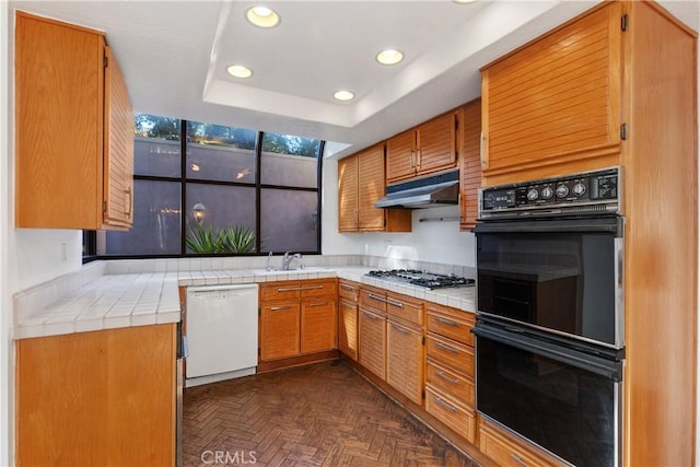 kitchen with white dishwasher, black double oven, a tray ceiling, tile counters, and stainless steel gas cooktop