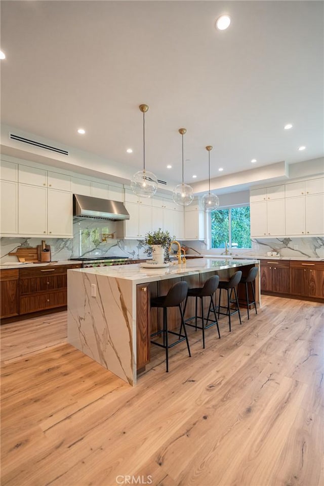 kitchen featuring light wood-type flooring, ventilation hood, a large island with sink, pendant lighting, and white cabinetry