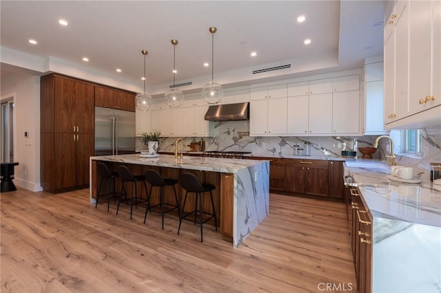 kitchen featuring a large island with sink, ventilation hood, light stone countertops, light wood-type flooring, and stainless steel appliances