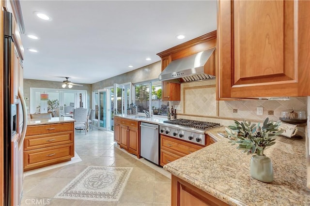 kitchen featuring wall chimney exhaust hood, ceiling fan, decorative backsplash, light stone countertops, and stainless steel appliances