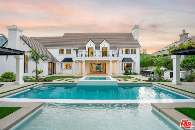 back house at dusk with a patio area, a balcony, and a pool with hot tub