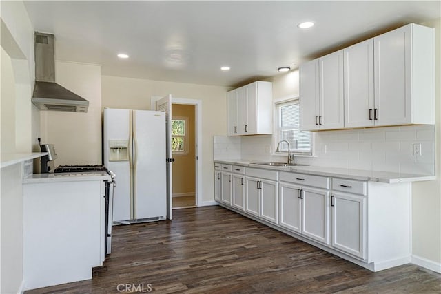 kitchen with white refrigerator with ice dispenser, light countertops, white cabinets, a sink, and wall chimney range hood