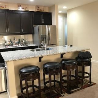 kitchen with a center island with sink, stainless steel refrigerator with ice dispenser, sink, light stone countertops, and a kitchen breakfast bar