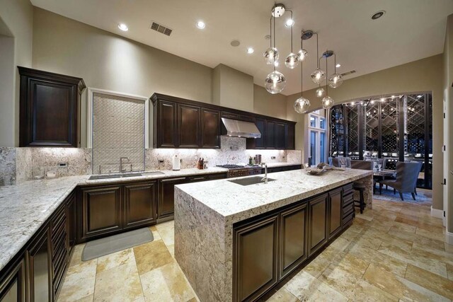 kitchen with decorative backsplash, wall chimney exhaust hood, a kitchen island with sink, sink, and decorative light fixtures