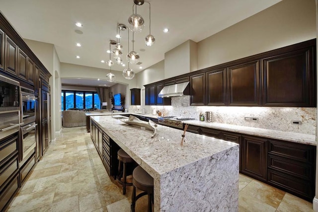 kitchen with a large island with sink, ventilation hood, decorative light fixtures, light stone counters, and a breakfast bar area