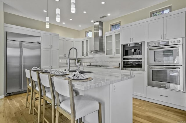 kitchen with pendant lighting, wall chimney range hood, appliances with stainless steel finishes, a kitchen island with sink, and light stone countertops