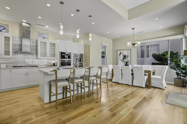 kitchen featuring black stovetop, an island with sink, pendant lighting, wall chimney range hood, and white cabinets