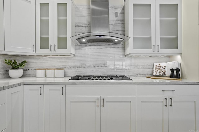 kitchen with wall chimney exhaust hood, stainless steel gas cooktop, white cabinetry, light stone counters, and backsplash