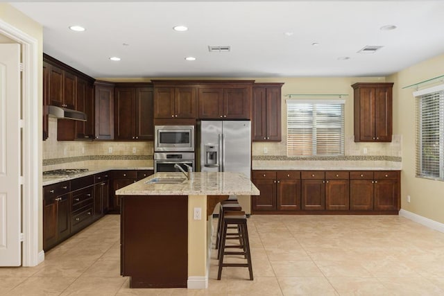 kitchen with backsplash, a center island with sink, a breakfast bar area, light stone counters, and stainless steel appliances