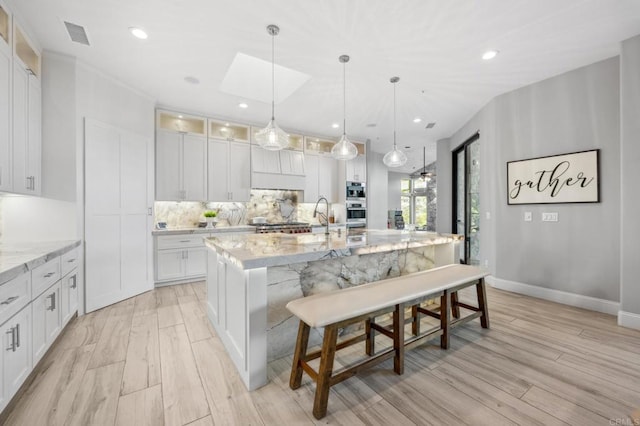 kitchen with white cabinetry, light stone counters, light hardwood / wood-style flooring, pendant lighting, and a center island with sink