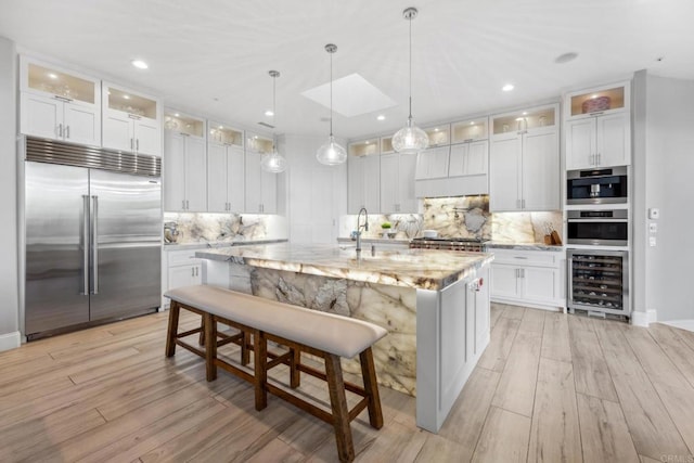 kitchen with white cabinets, wine cooler, an island with sink, light stone counters, and stainless steel appliances