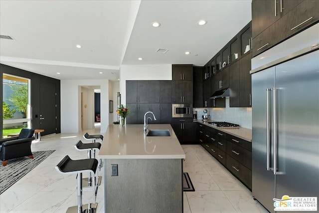 kitchen featuring a kitchen bar, backsplash, a kitchen island with sink, built in appliances, and sink