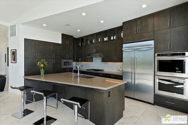 kitchen featuring tasteful backsplash, sink, built in appliances, dark brown cabinetry, and a breakfast bar area