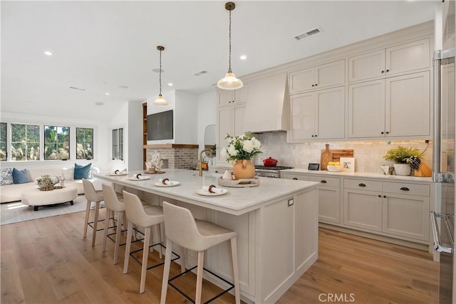 kitchen featuring a center island with sink, decorative light fixtures, custom range hood, and light hardwood / wood-style floors