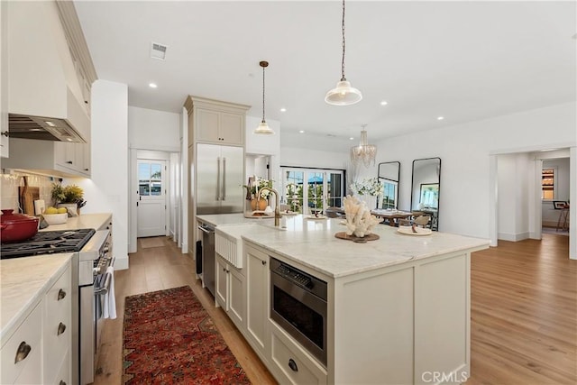 kitchen with hanging light fixtures, light hardwood / wood-style flooring, light stone countertops, an island with sink, and appliances with stainless steel finishes