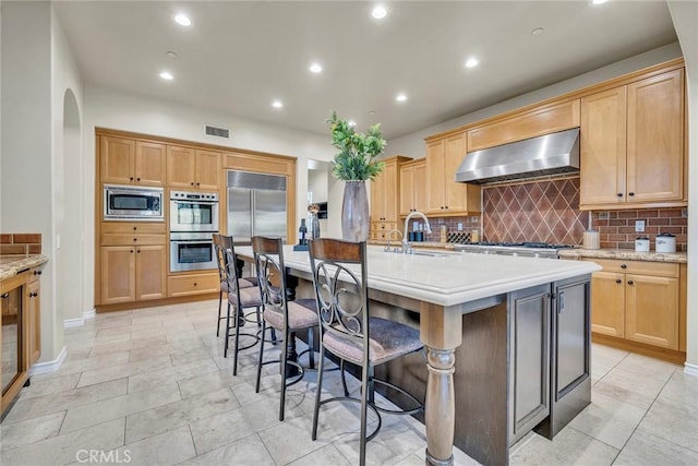 kitchen featuring backsplash, a center island with sink, sink, built in appliances, and wall chimney exhaust hood