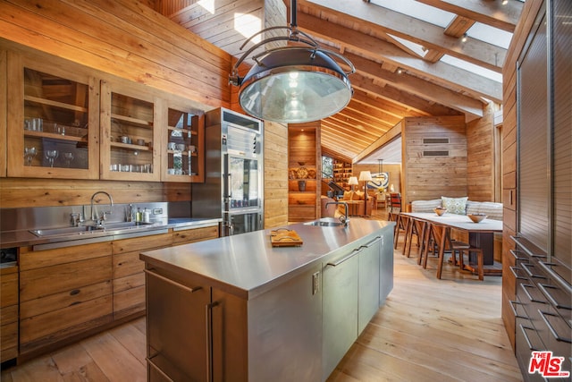 kitchen with wood walls, lofted ceiling with skylight, sink, light hardwood / wood-style flooring, and an island with sink