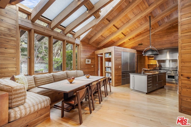 dining space with a skylight, beam ceiling, high vaulted ceiling, light hardwood / wood-style floors, and wood walls