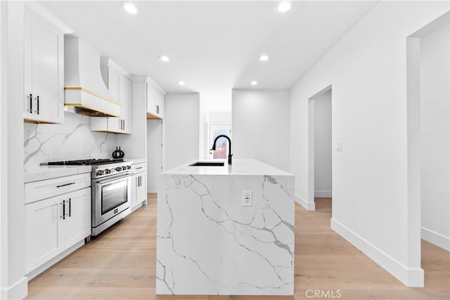 kitchen featuring custom exhaust hood, white cabinets, stainless steel stove, light wood-type flooring, and an island with sink